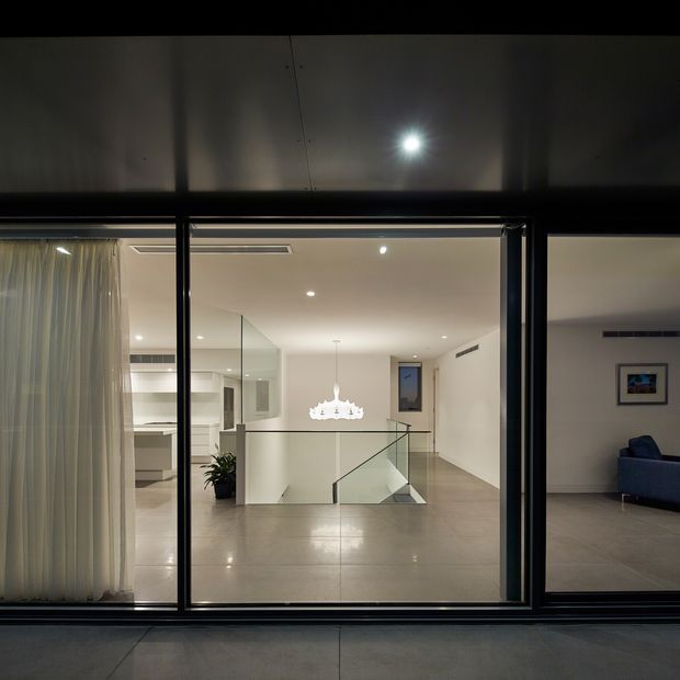 View through large black-framed glazing into a contemporary multi-level interior. The scene centres on a tiled landing with a frameless glass balustrade and stair opening, leading toward a bright open-plan kitchen and dining area with a sculptural pendant light. A living room with a blue sofa is visible to the right, while sheer curtains and reflective glazing frame the foreground. The space has a restrained neutral palette and clean, minimalist detailing.