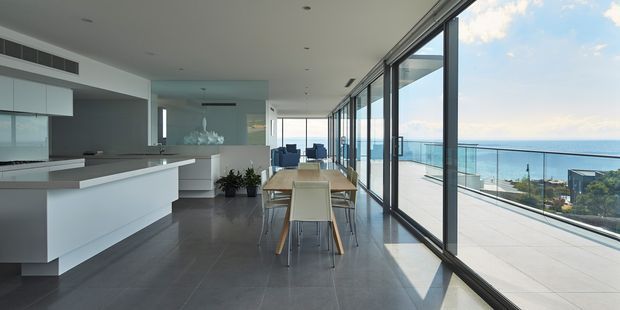 A bright, open-plan contemporary interior combining a sleek white kitchen, dining area, and living space. Floor-to-ceiling glazed sliding doors run along the right side, opening to a spacious terrace with expansive ocean views. The room has polished grey flooring, minimalist white cabinetry, a timber dining table with light upholstered chairs, and strong natural light.