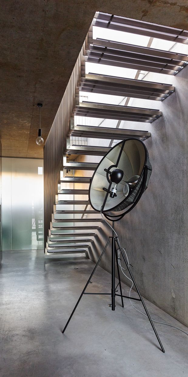 Industrial-style interior corridor or stair hall with a raw concrete floor, ceiling and wall, exposed steel stair structure, and a long run of open-riser stairs beneath a linear skylight. A large black pedestal fan stands in the foreground, while frosted glass doors and dark vertical wall panelling are visible at the far end. The image has a minimalist, utilitarian architectural character with strong natural light from above.
