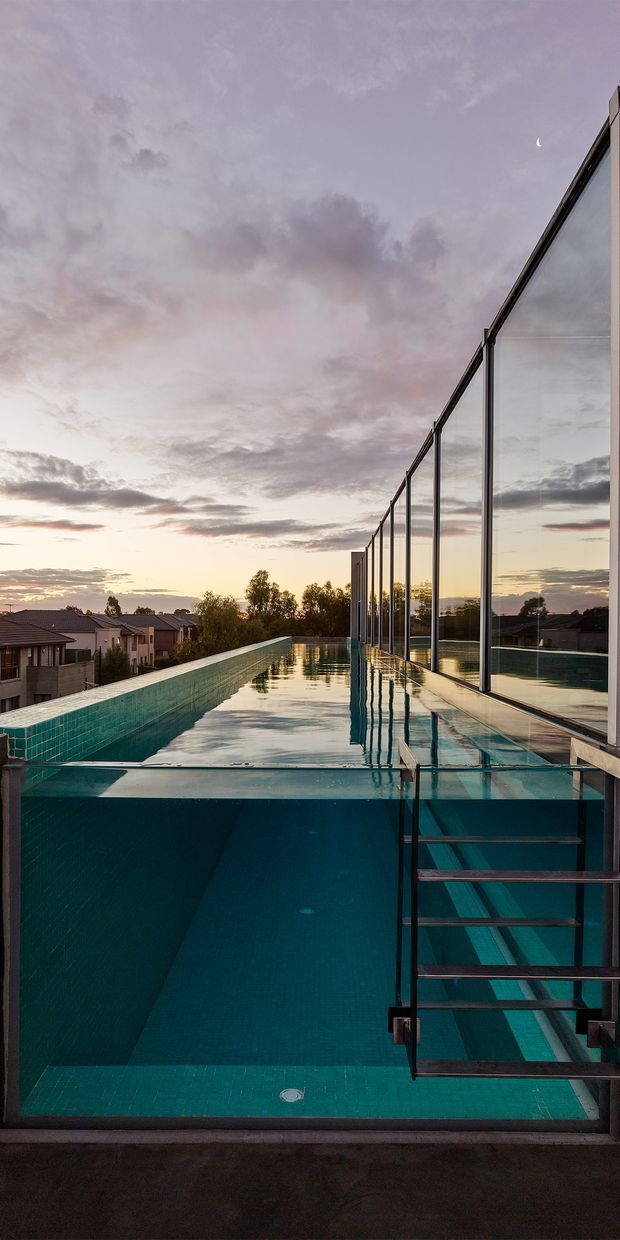 A dramatic dusk view of a long, narrow elevated swimming pool beside a modern building. The pool has deep turquoise tiled walls, a glass edge, and a metal staircase descending into the water. Full-height glazing runs along the right side, reflecting the pool and sunset sky, while a suburban neighbourhood is visible beyond the pool.