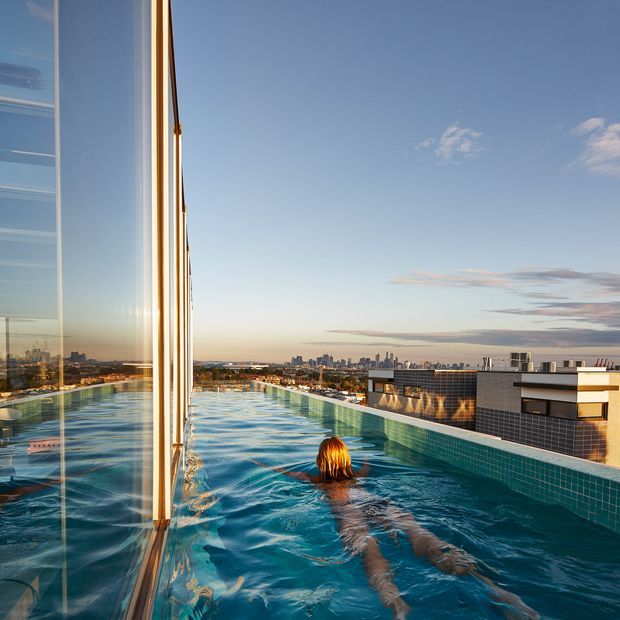 Rooftop infinity pool beside floor-to-ceiling glazing, with a swimmer in the water and a city skyline visible beyond. The pool is lined with small turquoise-blue tiles and bordered by a tiled parapet, creating a modern urban rooftop setting in warm late-afternoon light.