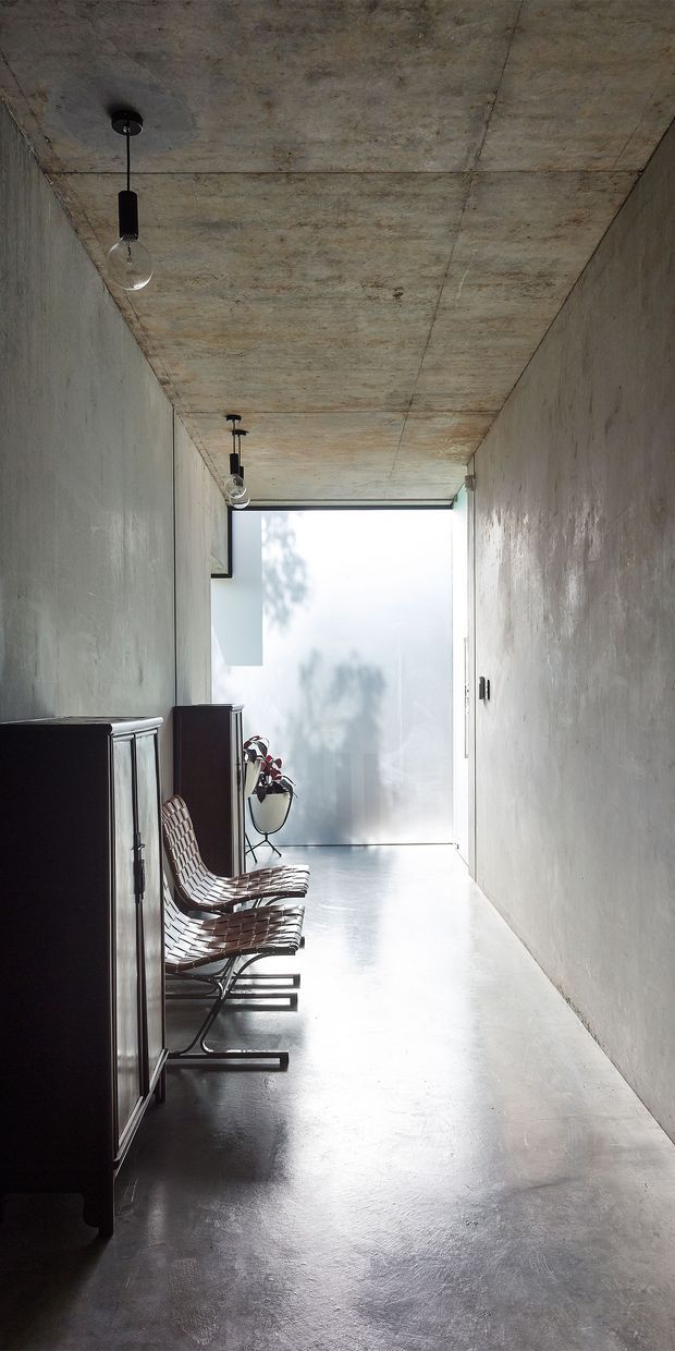 A narrow, minimalist concrete hallway or passage with raw poured-concrete walls and ceiling, a smooth grey polished floor, exposed black pendant bulbs, dark built-in cabinetry, and two woven leather sling chairs. Bright frosted glazing at the far end creates a calm, gallery-like atmosphere.