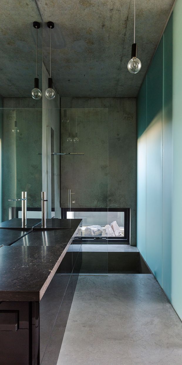 A narrow, sophisticated bathroom with a long dark stone vanity and integrated basin, twin polished metal taps, full-height mirrors and glass shower screening. The room features a raw mottled concrete ceiling and walls, polished concrete flooring, dark built-in cabinetry, slim black-framed glazing, and turquoise-blue wall panelling. Three suspended globe pendant lights provide a distinctive architectural feature, while natural light enters from the side window.