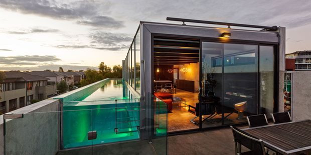 Rooftop terrace at dusk with a transparent-edge swimming pool beside a compact glazed pavilion. The pavilion has dark metal framing, extensive glass sliding doors, exposed ceiling beams, warm interior lighting, and a small lounge/dining area. A dark timber-look outdoor dining table sits in the foreground, with neighbouring low-rise buildings visible beyond.