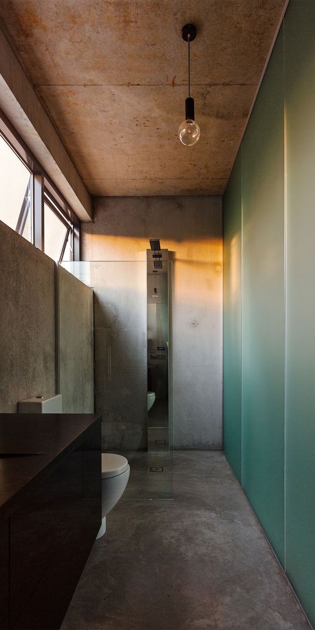 A narrow contemporary bathroom with an industrial aesthetic. The space features a raw concrete ceiling, concrete-look walls and floor, a frameless glass shower enclosure with a slim shower column, a white toilet, and dark built-in vanity cabinetry. Tall windows bring in natural light, while a teal-green wall and a simple exposed-bulb pendant add colour and character.