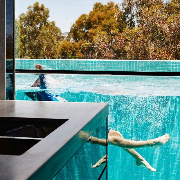 A close-up view from an outdoor kitchen or entertaining area looking through a large glazed pool wall into a turquoise swimming pool. A swimmer is visible underwater, with turquoise mosaic pool tiling, dark framing, and a pale stone benchtop in the foreground. Mature trees are visible beyond the pool, creating a striking indoor-outdoor connection.