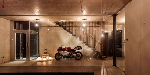 A warmly lit, industrial-style interior featuring polished concrete walls and ceiling, a glass-enclosed lift or display enclosure on the left, and a sculptural open-riser staircase with slender metal balustrade. A motorcycle is prominently displayed behind full-height glazing, with built-in shelving and concealed lighting adding to the gallery-like atmosphere.