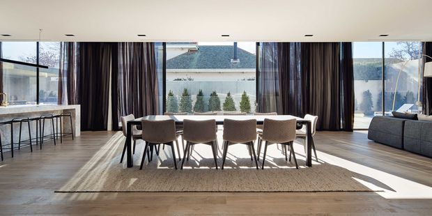 A bright, spacious open-plan dining and living area with a long rectangular dining table and upholstered chairs, expansive floor-to-ceiling glazing, dark full-height curtains, and a large sectional sofa. A kitchen island with bar stools is visible at left. Warm timber flooring and abundant natural light create a refined contemporary interior with views toward neighbouring homes and a distant landscape.