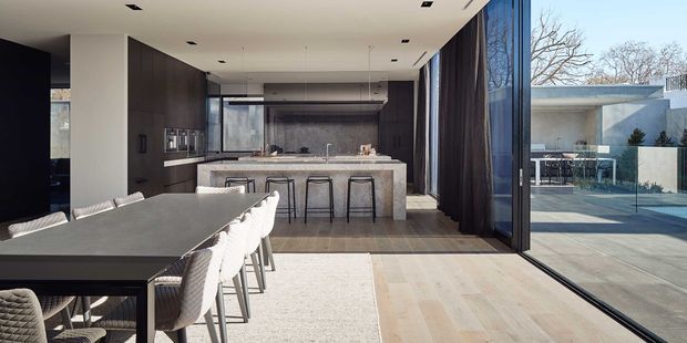A refined open-plan dining and kitchen interior with a long dark dining table and upholstered chairs in the foreground, leading to a large stone kitchen island with bar stools. Dark timber cabinetry, pale timber flooring, recessed ceiling lights, and full-height glazed sliding doors create a sleek connection to an outdoor terrace and pool area.