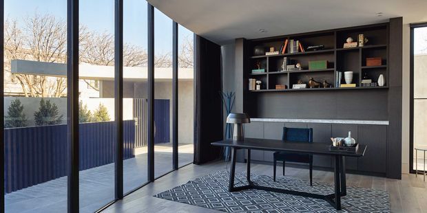 A refined contemporary home office with a dark built-in shelving and cabinetry wall, integrated desk, and floor-to-ceiling black-framed glazing. The room opens visually to a paved courtyard, with a patterned rug, desk accessories, books, and restrained decor creating a sophisticated, moody workspace.