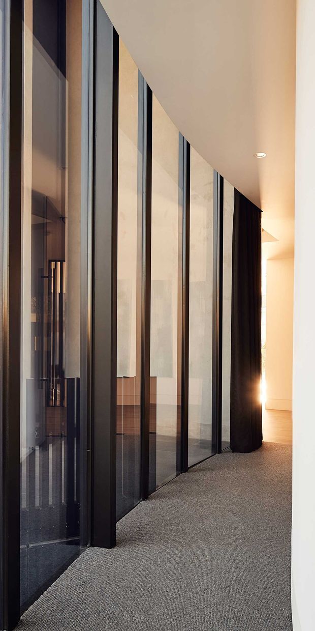 A narrow, softly lit interior corridor with a sweeping curved wall of floor-to-ceiling black-framed glass partitions. Dark curtains, pale plaster walls, recessed ceiling lighting, and a grey carpet create a refined, contemporary architectural interior. Reflections reveal additional interior elements behind the glazing.