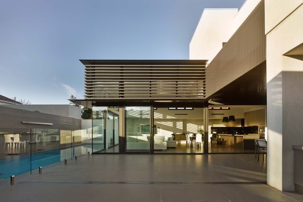 Contemporary multi-level residential exterior opening onto a broad paved terrace, with extensive floor-to-ceiling glazing, a slatted horizontal pergola or brise-soleil, and a visible open-plan living and kitchen area beyond. A swimming pool and glass balustrade sit to the left, while clean white rendered walls and dark framed glazing create a crisp indoor-outdoor connection.