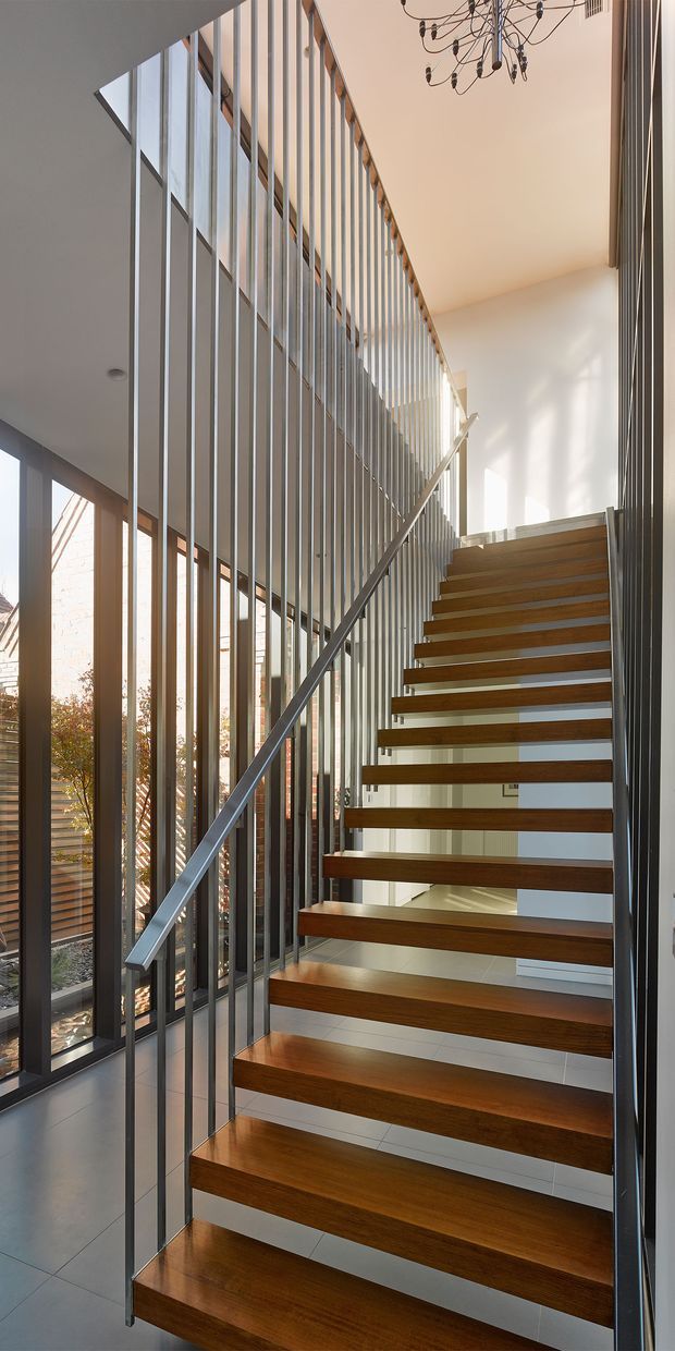 A tall, light-filled contemporary stairway with floating dark timber treads, slim steel stringers and handrail, and a full-height vertical metal balustrade forming a striking screen. Floor-to-ceiling glazing with timber-framed windows lines the stairwell, while a sculptural branching pendant hangs from the high ceiling. The space has a refined, minimalist architectural character with strong indoor-outdoor visual connection.