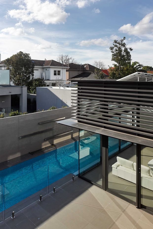 Elevated view of a contemporary residential courtyard with a vivid blue swimming pool, dark tiled terrace, frameless glass pool balustrade, and large black-framed glazed doors. A horizontal slatted privacy screen shades the upper level, with neighbouring suburban homes and mature trees visible beyond.