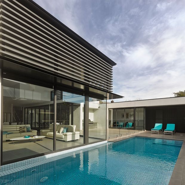 Contemporary multi-level residence viewed from the pool courtyard, featuring extensive floor-to-ceiling glazing, a projecting upper level screened with horizontal fins, dark-framed windows, pale rendered walls, and a vivid blue tiled swimming pool. The glass facade reveals a refined living room with sofas and a low coffee table, while a covered outdoor area and turquoise pool loungers occupy the opposite side. The image has a clean, high-contrast architectural photography style with strong geometric lines.