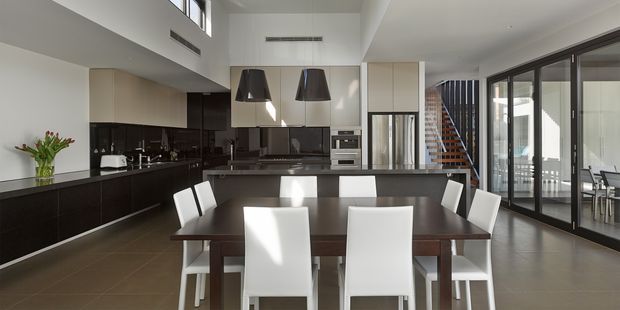 A spacious open-plan kitchen and dining area in a contemporary home. The kitchen features dark glossy lower cabinetry, pale upper cabinetry, dark reflective splashback surfaces, integrated stainless-steel appliances, and a central island with two black pendant lights. A large dark wood dining table with white upholstered chairs occupies the foreground. Full-height black-framed glazed doors open toward an outdoor area, while a staircase and double-height volume are visible in the background. The palette is monochromatic and refined with strong natural daylight.