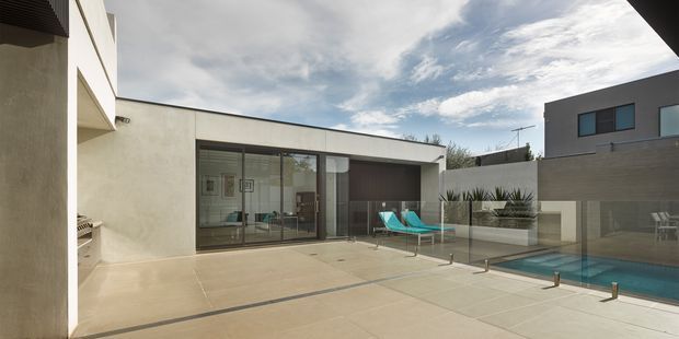 A contemporary flat-roofed courtyard residence viewed from a broad paved terrace. The composition features pale rendered walls, extensive dark-framed glazing, a raised rectangular pool, built-in planters with spiky ornamental plants, and a neighbouring upper-level structure. Turquoise outdoor loungers provide a small colour accent against the restrained neutral architecture.