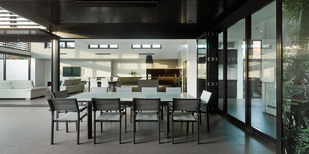 A spacious contemporary open-plan living and dining area with a long dark dining table and multiple black-framed chairs in the foreground. Floor-to-ceiling glazed sliding doors line the right side, opening toward an exterior garden or courtyard. A bright lounge with white sofas sits to the left, while a sleek kitchen with dark cabinetry and a central island is visible at the rear. The room has a dark slatted ceiling, pale tiled flooring, extensive glazing, and strong natural daylight.