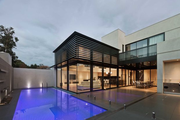A contemporary multi-level residence viewed from an outdoor pool terrace at dusk. The house features extensive floor-to-ceiling glazing, a projecting upper volume screened by horizontal dark slats, pale rendered walls, dark metal-framed windows and doors, and an outdoor dining area beside an illuminated swimming pool.