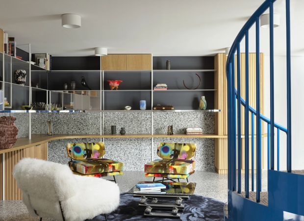 A bright, eclectic living room with a large built-in shelving and storage wall, featuring dark open shelving, timber cabinetry, terrazzo-like speckled panels, and decorative objects. Two colourful sculptural lounge chairs sit around a low glass coffee table on a dark patterned rug, with a white sheepskin-covered chair in the foreground. A distinctive blue metal spiral staircase railing occupies the right side of the room.