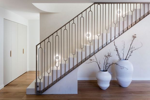 Bright contemporary stairway with a sculptural dark metal balustrade featuring repeated rounded vertical motifs. White walls, concealed white storage cabinetry, warm timber flooring, and large white ceramic vessels with bare branches create a clean, refined interior.
