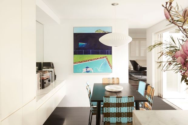 Bright contemporary dining area with a dark rectangular dining table and mixed turquoise woven chairs, positioned beside full-height glazing and a pale stone island or counter. White built-in cabinetry and shelving line the left wall, while a sculptural white pendant light hangs above the table. A colourful abstract artwork provides a strong accent, and a glimpse of an adjoining lounge or study is visible beyond.