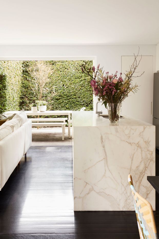 Bright contemporary open-plan kitchen and living area with a large white marble waterfall island, dark timber flooring, white cabinetry, and an adjacent outdoor terrace visible through wide glazing. A sculptural floral arrangement sits on the island, while white seating and minimalist outdoor furniture reinforce the airy, refined aesthetic.
