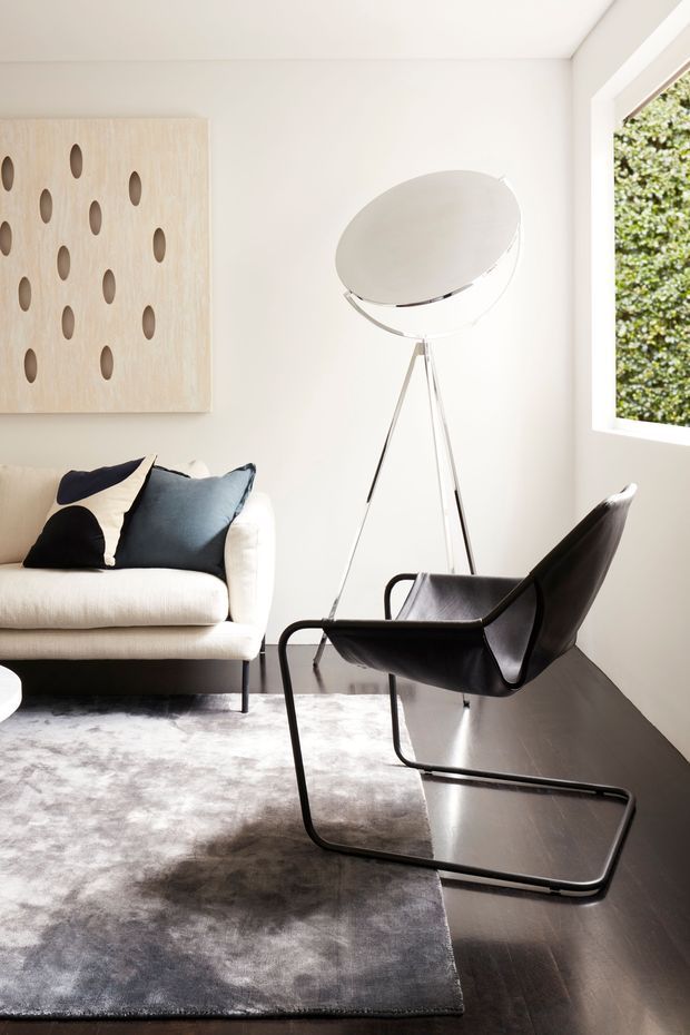 Bright, refined living room with a cream upholstered sofa, sculptural black leather lounge chair, tripod floor lamp, pale perforated wood wall artwork, grey rug, and dark timber flooring. Large window provides natural light and greenery beyond.