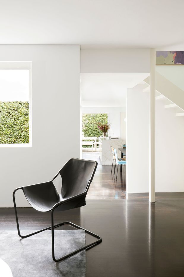 Bright minimalist interior view featuring a sculptural black leather lounge chair on a pale rug, smooth white walls, dark timber flooring, and an open sightline into a dining and kitchen area. White structural columns, a stairway, large windows, and garden views create a clean, airy composition.