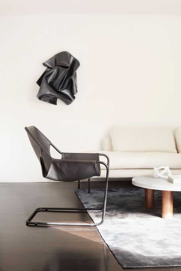 A refined living room with a white upholstered sofa, sculptural black leather lounge chair, round pale stone coffee table, dark timber flooring, and a large grey rug. A black abstract wall sculpture provides a dramatic focal point against the bright white wall.