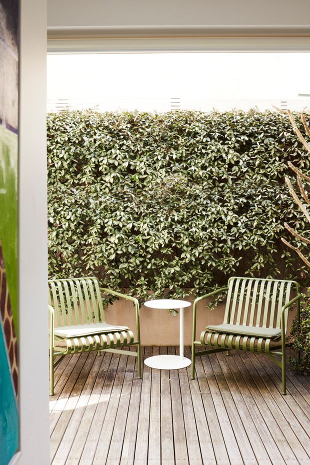 A compact sheltered outdoor courtyard or deck viewed through a doorway, with two pale green slatted lounge chairs and a small white round side table on weathered timber decking. A dense clipped hedge forms the privacy backdrop, with a simple overhead covering and partial interior wall framing the scene.