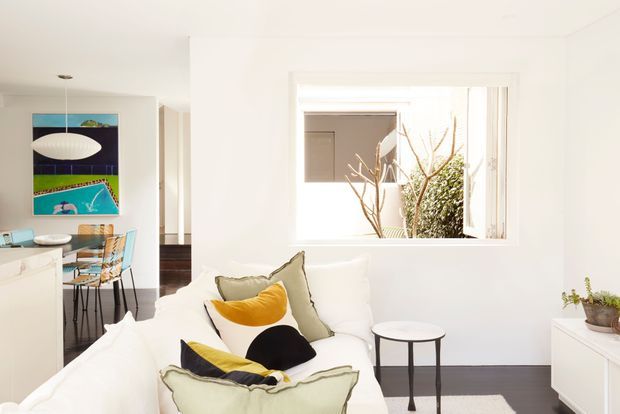 Bright, compact contemporary living area with a white sofa, layered cushions, a small round side table, and a low white storage unit. An open-plan kitchen and dining area is visible to the left, with a pendant light, bar seating, and colourful wall art. White walls, dark timber flooring, and a large internal window create an airy, high-contrast interior.