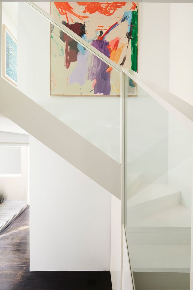 Bright minimalist interior stairway with white painted walls, white stair treads, and a clear glass balustrade framed in white. An abstract multicolour artwork hangs above the stairs, while dark timber flooring extends into the adjoining space.