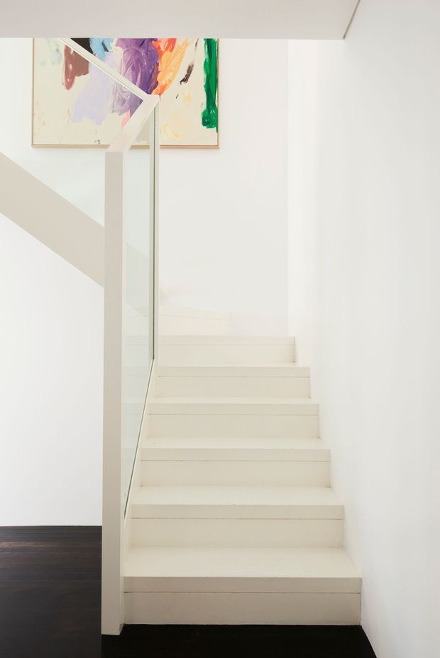 Minimalist interior stairway with white painted walls and a white staircase, clear glass balustrade, and a dark timber floor. Abstract colourful artwork is mounted near the upper landing, providing a vivid accent against the otherwise monochromatic space.