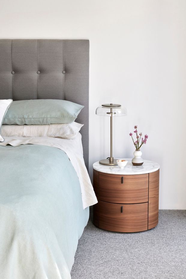 A softly styled bedroom vignette featuring a bed with a tall grey upholstered, button-tufted headboard, pale blue-green and white bedding, and a rounded walnut bedside table with a white marble top. The table holds a slim brass-and-glass lamp and a small ceramic vase with flowers. Light grey carpet and white walls create a calm, refined interior.