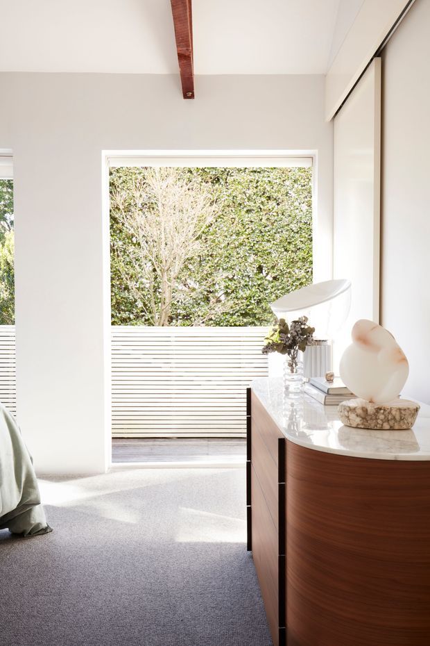 Bright, refined bedroom interior with a large framed window opening to a leafy outdoor area and slatted privacy screen. The room features pale walls and carpet, a warm timber ceiling beam, and a curved timber dresser with a glossy light benchtop. Minimal decorative styling includes a sculptural lamp, vase, books, and organic ornament.