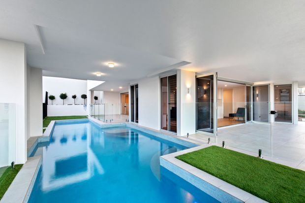 Modern covered outdoor pool courtyard with bright blue swimming pool, white rendered walls, pale tiled paving, glass balustrades, large glazed doors, and small manicured lawn sections. The space has a clean, resort-like indoor-outdoor design with recessed lighting and contemporary detailing.