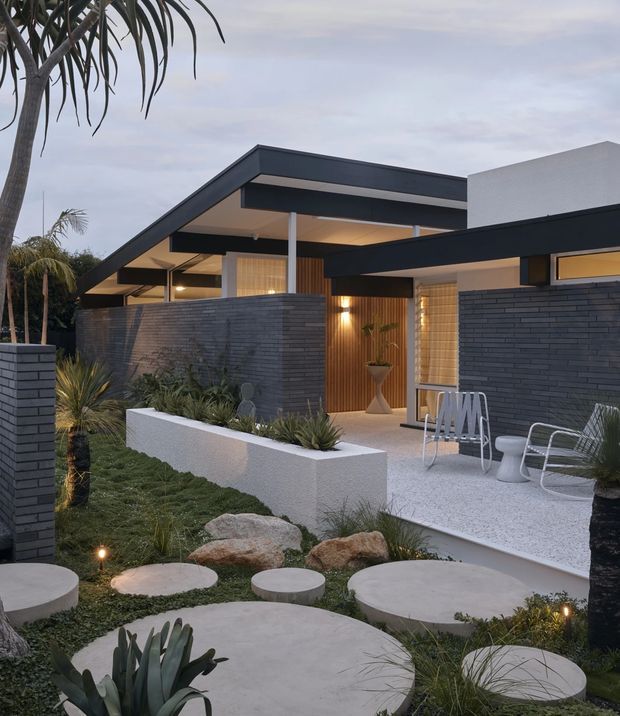 Contemporary single-storey residence with strong horizontal flat-roof forms, dark charcoal brick walls, white rendered volumes, warm timber entry detailing, and expansive outdoor landscaping. The front garden features sculptural concrete stepping pads, native-style grasses and succulents, boulders, low planting beds, and a small paved outdoor seating area with modern metal chairs. Soft exterior lighting creates a warm evening atmosphere.