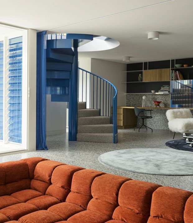 A bright open-plan living room with a bold blue spiral staircase, terrazzo flooring, and a built-in study and kitchen area at the rear. The space features a large rust-orange modular sofa in the foreground, a round pale rug, blue curtains, a white textured armchair, dark shelving, and a curved mezzanine or stair opening. Floor-to-ceiling glazing brings in abundant daylight.