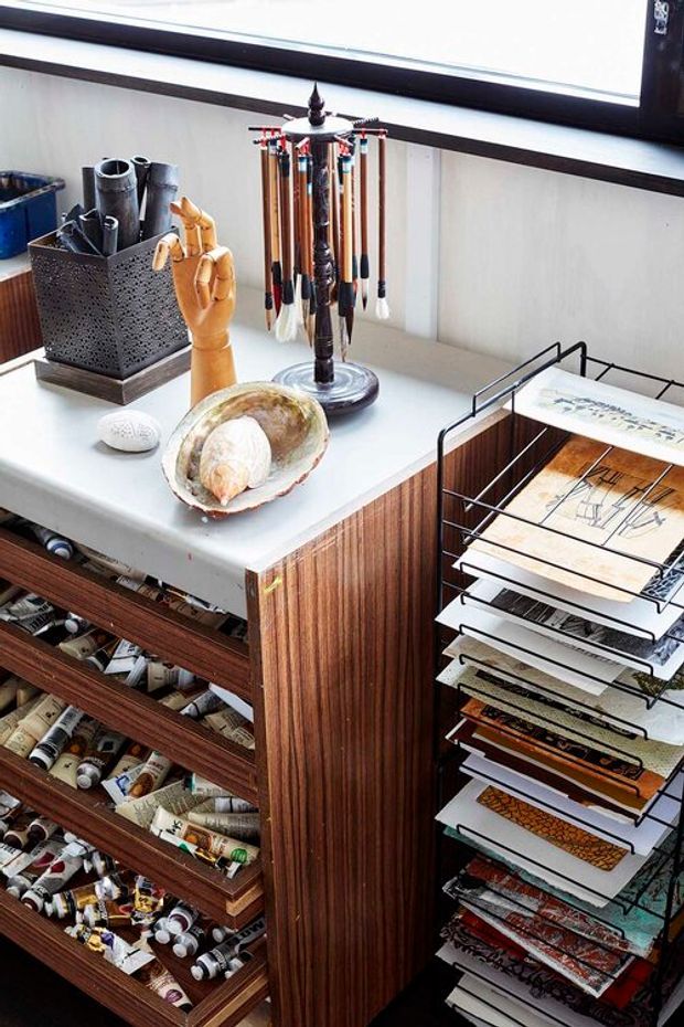 Close-up of a compact, highly organized creative work station beneath a large window. A white worktop sits over dark, vertically grained timber cabinetry with multiple open drawers containing art supplies, while a freestanding metal document rack holds papers and prints. The surface includes a pen or brush carousel, a perforated container of tools, a wooden mannequin hand, and decorative shells. The scene has a functional studio or home-office character with strong mid-century-inspired timber joinery.