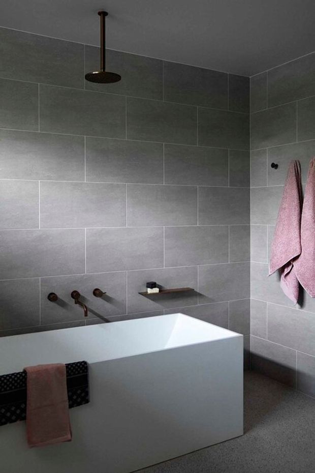 A compact contemporary bathroom featuring a freestanding white bathtub, large-format grey rectangular wall tiles, a dark bronze ceiling-mounted rain shower, matching bronze bath fixtures, and soft pink towels. The room has a restrained, moody palette with minimalist built-in detailing.