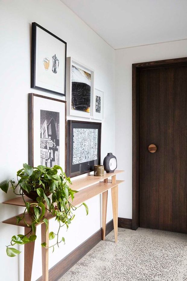 Bright, minimalist entryway featuring a slim light-timber console with open shelves, framed monochrome artwork, a leafy indoor plant, and a large dark timber door with a round inset handle. White walls and a speckled terrazzo-like floor create a clean, high-contrast contemporary composition.