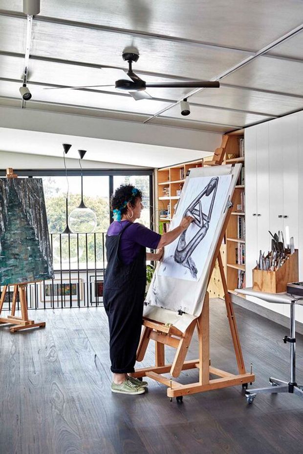 A bright contemporary artist's studio with a large easel and work-in-progress drawing in the foreground. Built-in timber shelving and white cabinetry line the right wall, with art supplies stored in a wooden container. Large glazing opens to a balcony or railing beyond, while a sculptural pendant light and a dark ceiling fan hang above. The room has a polished dark timber floor and an airy, functional creative-workspace atmosphere.