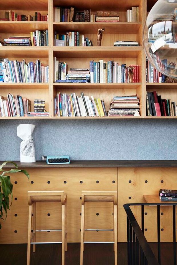 A compact study or library nook with extensive built-in timber shelving filled with books, a dark horizontal worktop, and a textured blue-grey backsplash or wall surface. Two timber stools sit at a perforated timber-fronted counter, with a small turquoise typewriter and decorative objects on the desk. A black stair or railing structure is visible at the right, and a hanging reflective globe light partially enters the upper-right corner. The space has warm timber joinery, dark flooring, and a curated, functional character.