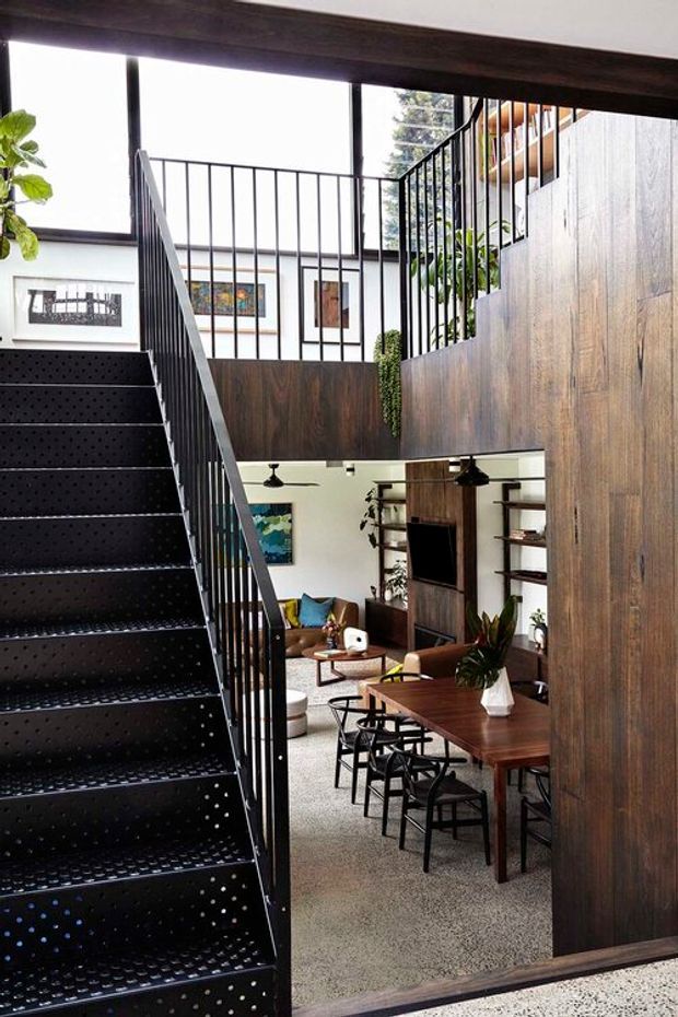 A contemporary multi-level interior centered on a dramatic black perforated metal staircase with slim black balustrades. Dark timber wall panelling and a matching upper-level partition frame views into an open-plan dining and living area below, furnished with a long timber dining table, black chairs, a sofa, shelving, and a television. White walls, framed artwork, indoor plants, ceiling fans, and abundant natural light create a refined yet warm atmosphere.
