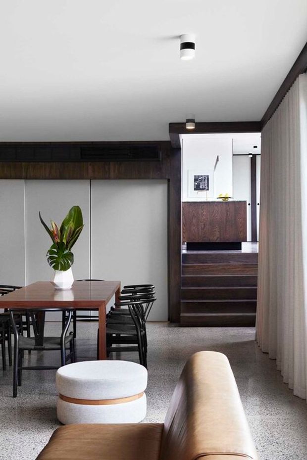 A refined open-plan dining and living area with a timber dining table and black chairs in the foreground, a low upholstered ottoman and leather sofa, and a compact stairway leading to a raised hallway or landing. The room features dark timber wall and stair joinery, pale built-in cabinetry, terrazzo-style flooring, a white ceiling with recessed spotlights, and full-height sheer curtains. The overall design is clean, warm, and contemporary with mid-century-inspired furniture.