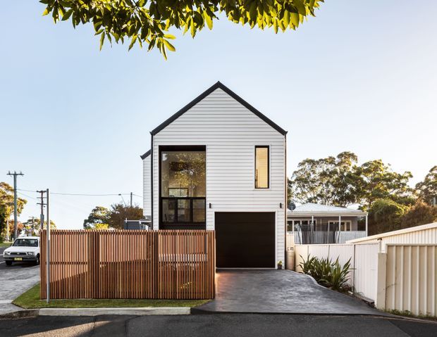 Street-facing view of a narrow two-storey residential house with a steep front gable, white horizontal weatherboard cladding, dark trim, a tall glazed entry, and an integrated ground-floor garage. A timber slat fence encloses the front setback and driveway, with neighbouring suburban homes and established trees around it.