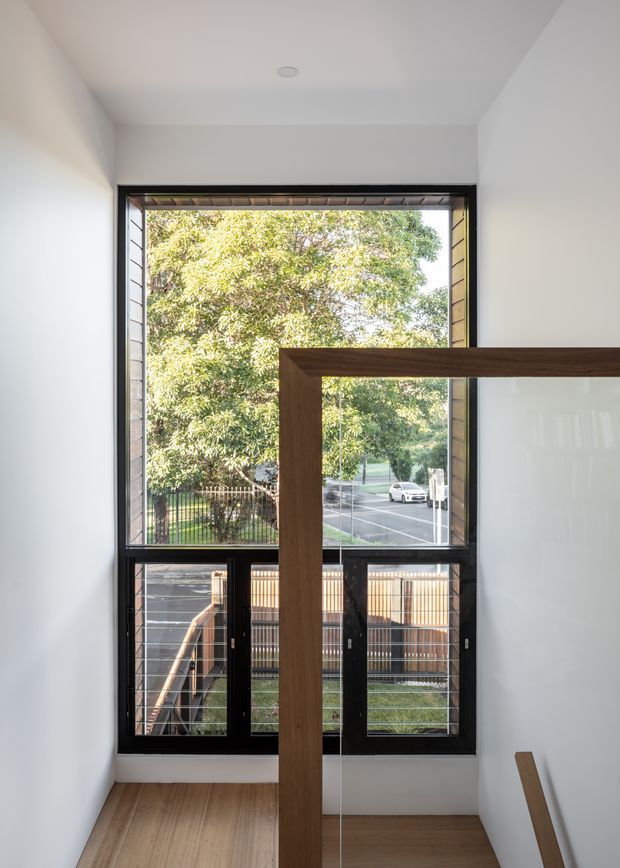 A bright, narrow interior stairway or landing with white walls, a light timber floor, and a large black-framed window overlooking trees, a street, and a neighbouring fence. A timber-framed glass balustrade runs through the foreground, emphasizing the verticality and openness of the space.