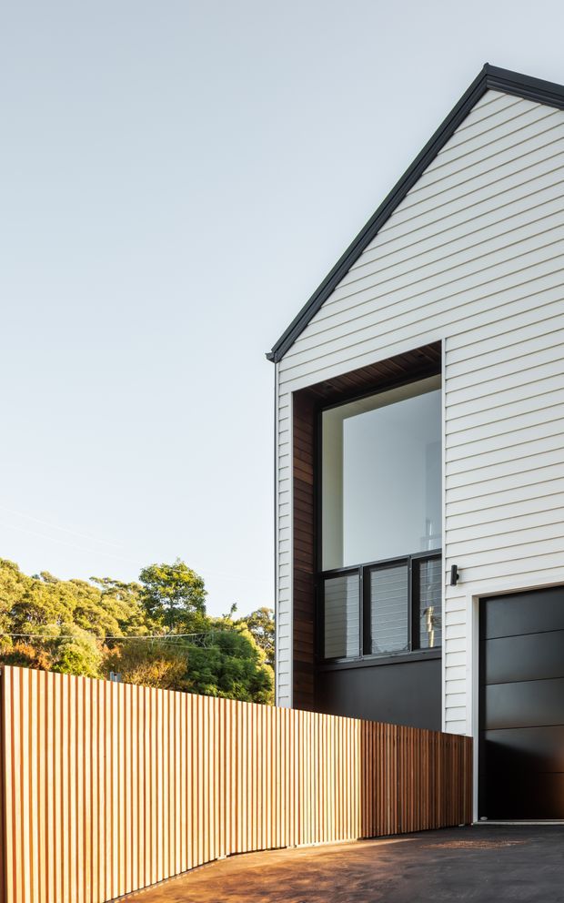 A contemporary multi-level residential exterior viewed from the driveway, featuring a steep gable roof, white horizontal weatherboard cladding, dark trim, a tall recessed balcony or entry void, dark garage door, and a warm timber slat privacy fence. Trees and open sky appear in the background.