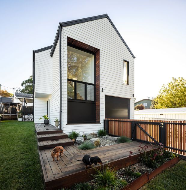 Exterior view of a compact contemporary two-storey home extension or townhouse, clad in pale horizontal weatherboards with dark trim and a steep gabled roof. A tall glazed entry opening is framed in dark timber, with a dark garage door below. The home opens onto a raised timber deck with timber fencing, landscaped planting beds, and steps from the lawn.
